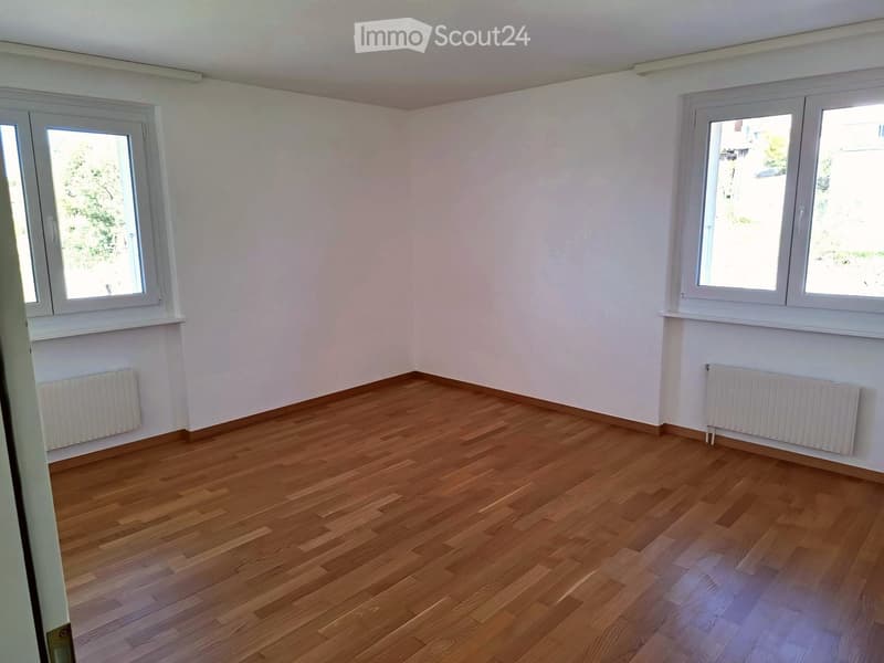 Empty room with wooden flooring, white walls, and large windows with white frames. Radiators are present under the windows on both sides.