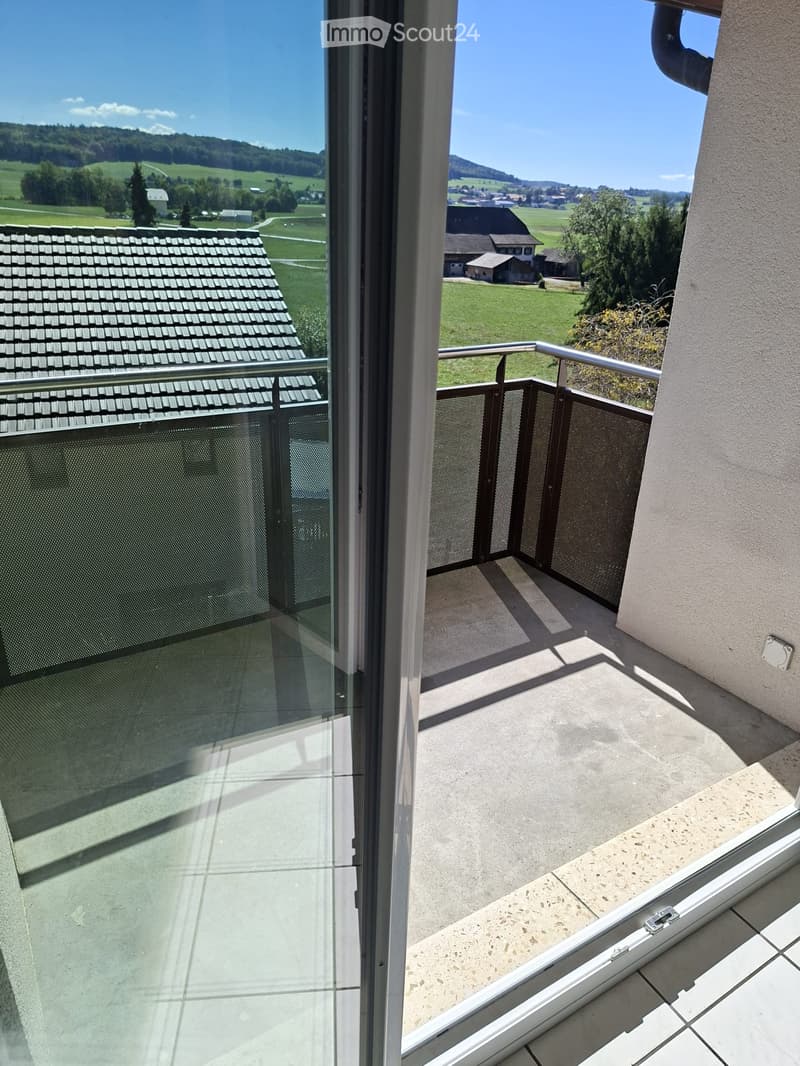 Balcony with glass railing, overlooking a green landscape with houses and hills.