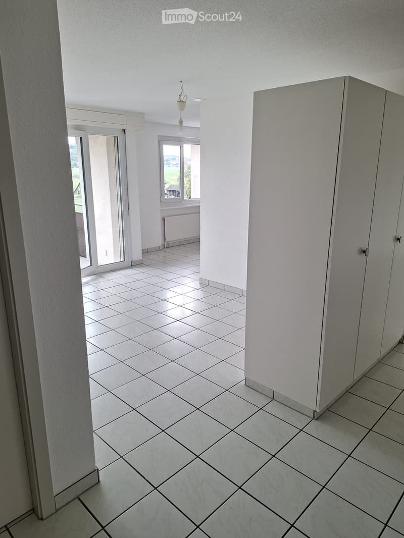 Empty hallway with white tiled flooring, white walls, and a hanging light fixture. Features a sliding glass door leading to an outdoor area and a window with a view of greenery.