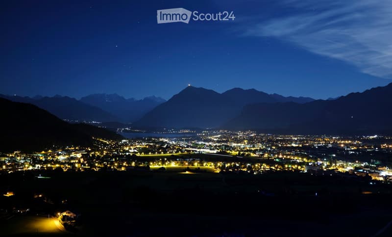 Nighttime view of a town with illuminated buildings and surrounding mountains under a starry sky.