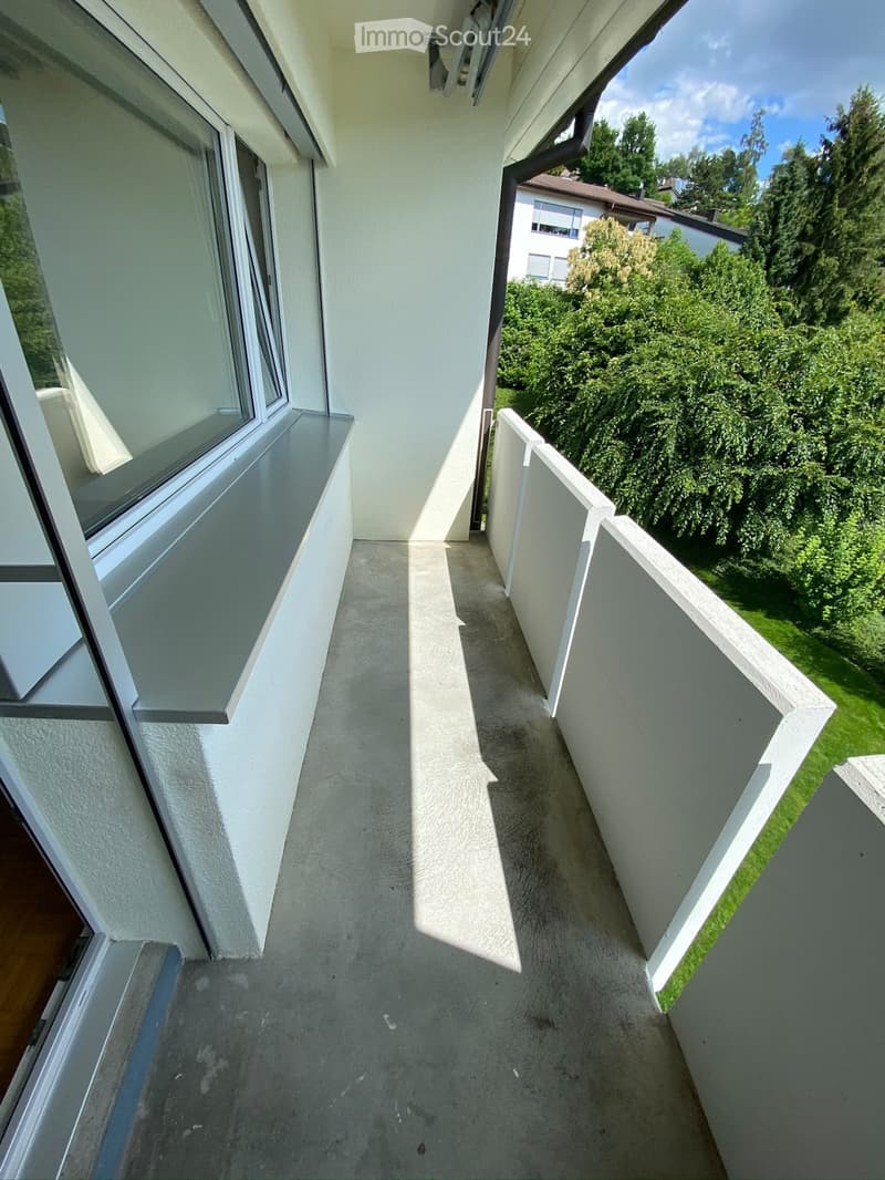 Balcony with white walls, concrete floor, and white railings, overlooking a green garden and neighboring building.