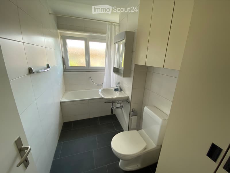 Modern bathroom with white tiles, bathtub, sink, toilet, mirror, and built-in cabinet. Features a window with frosted glass and a door with a silver handle.