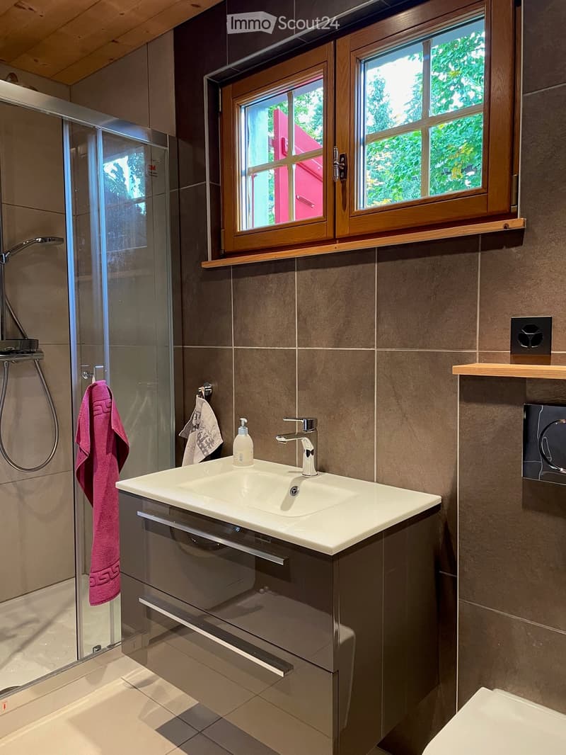 Modern bathroom with a white sink, chrome faucet, glass shower enclosure, and wooden window with a view of greenery.