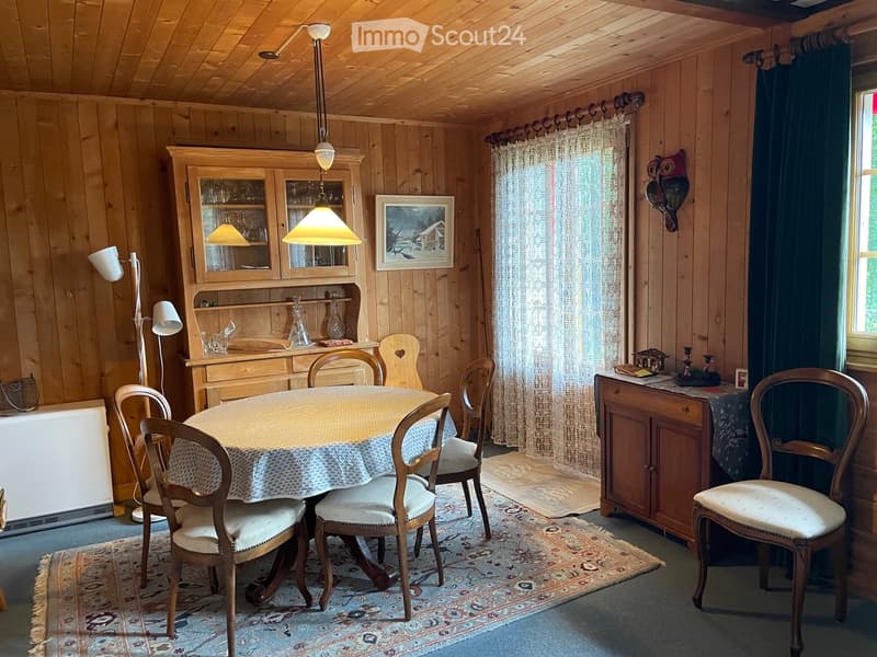 Cozy wooden-paneled dining room with a round dining table, six chairs, a wooden cabinet, and a sideboard. The room features a hanging pendant light, a floor lamp, and a decorative rug.