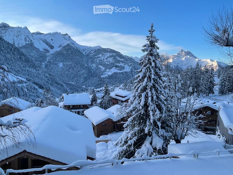Snow-covered village with wooden houses, snow-laden trees, and snow-capped mountains in the background.