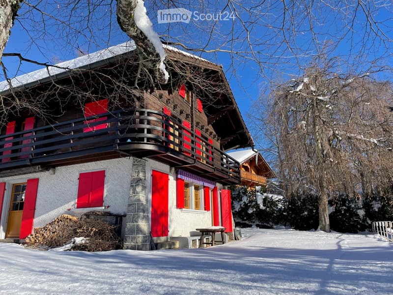 Two-story wooden house with red shutters, stone base, balcony, and snow-covered surroundings.