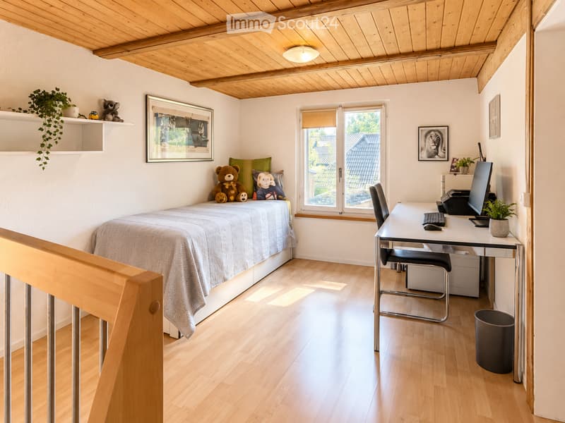 Cozy bedroom with a wooden bed, desk, chair, computer, and decorative items including a teddy bear and a plant. The room features light wooden flooring and white walls.