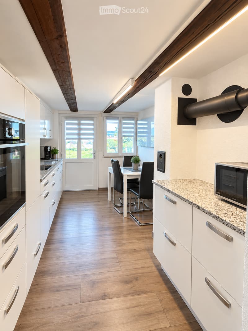 Modern kitchen with white cabinets, granite countertops, and a dining area with a table and chairs. Features include a microwave, coffee maker, and potted plants.