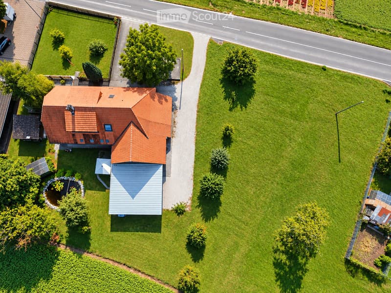 Aerial view of a two-story house with a red-tiled roof, surrounded by a well-maintained lawn, trees, and a driveway leading to a road.