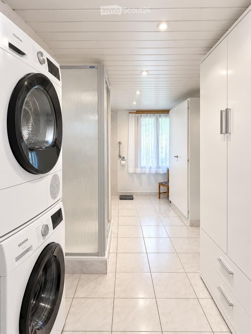 Clean, modern hallway with white tiled flooring, white cabinets, and a washing machine and dryer unit on the left side. A small bench and a window with white curtains are visible in the background.