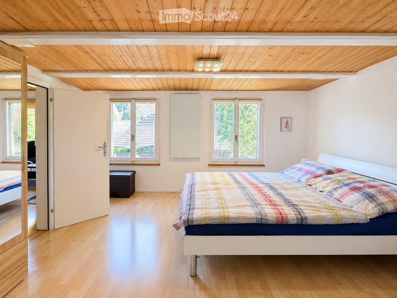 Bright bedroom with wooden floors and a slanted wooden ceiling. Features a bed with a colorful plaid duvet, a small black stool, and a picture frame on the wall. Large windows offer natural light and a view of the garden.