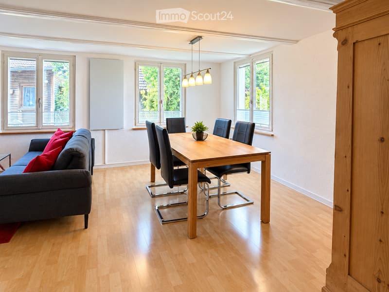 Bright living and dining area with hardwood flooring, large windows, a grey sofa with red cushions, a wooden dining table with four black chairs, and a small potted plant on the table.