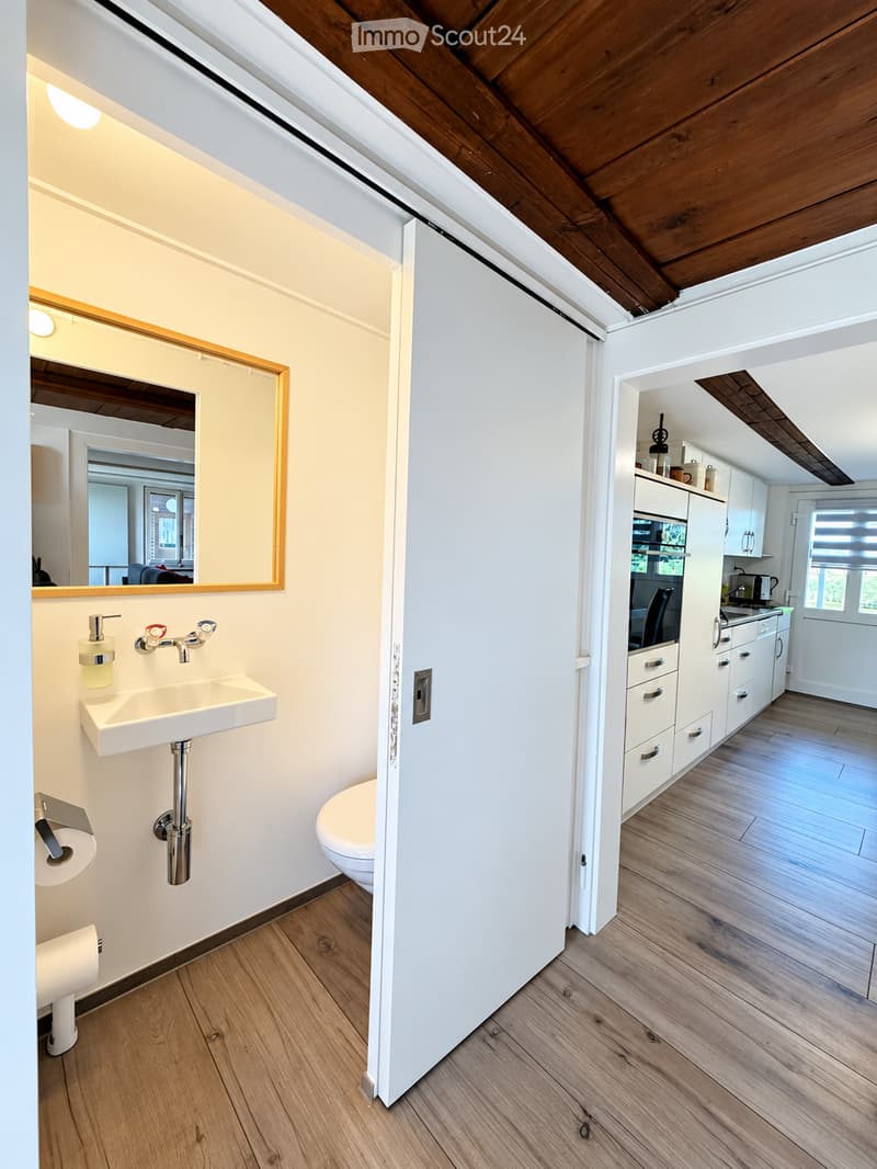 Modern bathroom with a white sink, toilet, and mirror. Adjacent kitchen area features white cabinets, an oven, and a microwave.