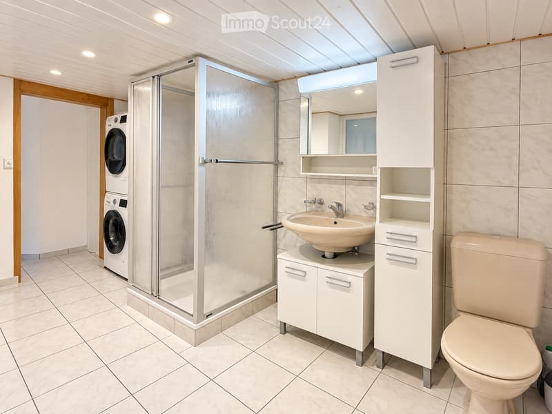 Modern bathroom with a white tiled floor, a glass-enclosed shower, a white sink with a cabinet, a toilet, and a washing machine in the background.