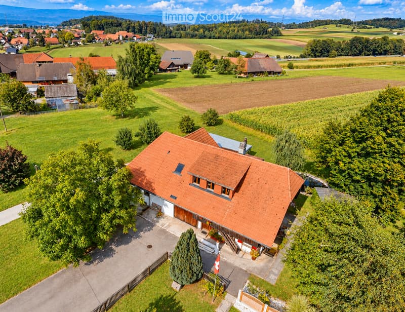 Aerial view of a two-story house with a red-tiled roof, surrounded by a well-maintained garden and a driveway. The house features multiple windows and a small balcony. The surrounding area includes green fields, trees, and neighboring houses.
