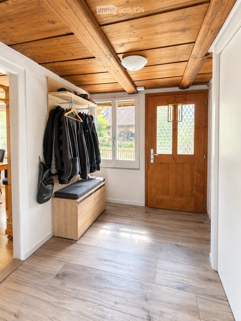 Bright hallway with wooden flooring, exposed wooden beams, built-in bench with storage, coat rack, and a large window offering a view of the garden.