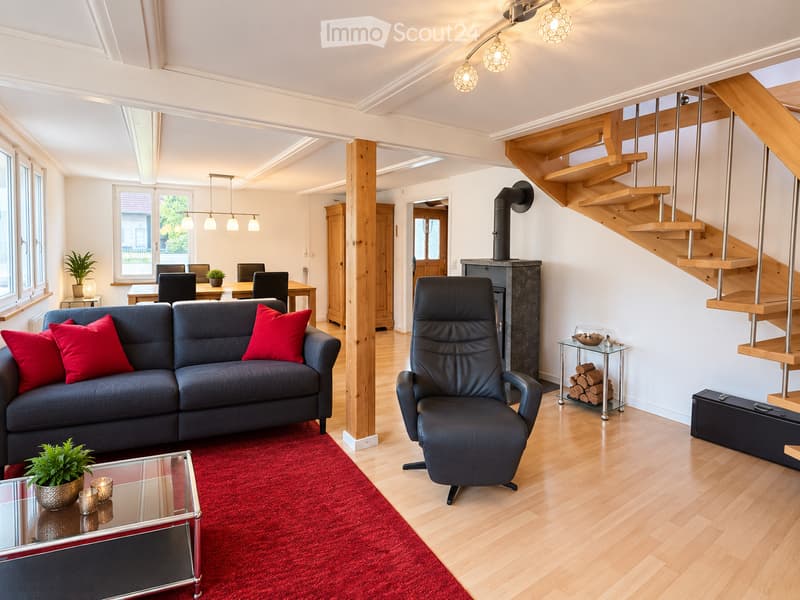 Modern living room with a grey sofa, red cushions, glass coffee table, armchair, dining area with table and chairs, fireplace, staircase, and potted plants.