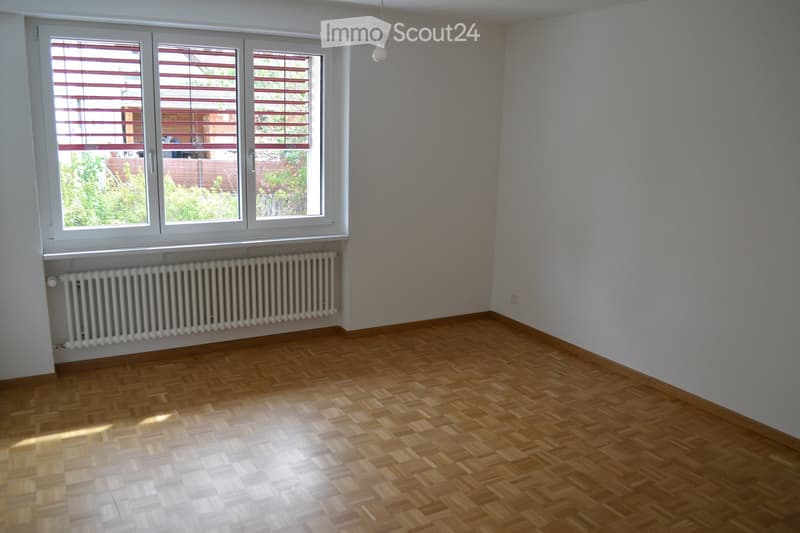 Empty room with wooden parquet flooring, white walls, large windows with blinds, radiator, and a hanging light fixture.