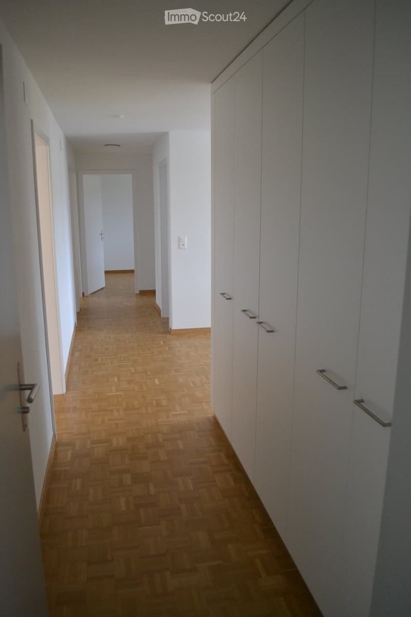 A long, narrow hallway with wooden flooring and white walls, featuring built-in cabinets with metal handles on one side and doors leading to other rooms.