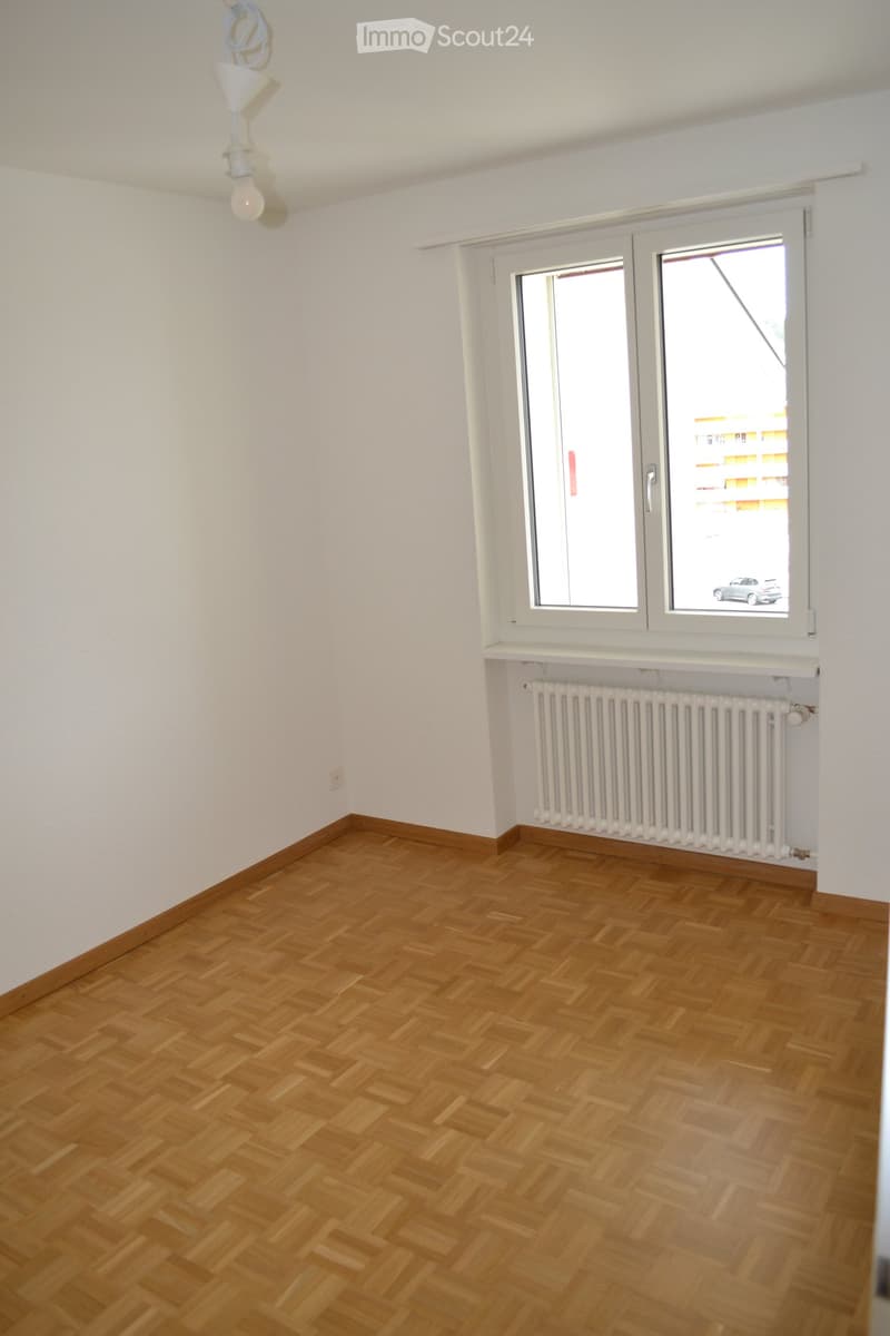 Empty room with wooden herringbone flooring, white walls, a radiator, and a large window with a view of a building and parked car.