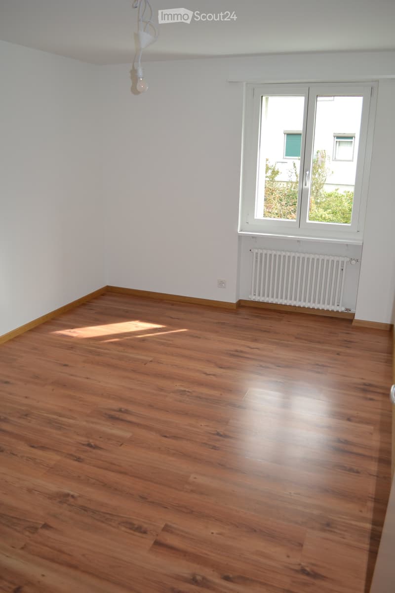 Empty room with wooden flooring, white walls, and a large window with a radiator below it. The room has a simple, clean design with no furniture or decorations.