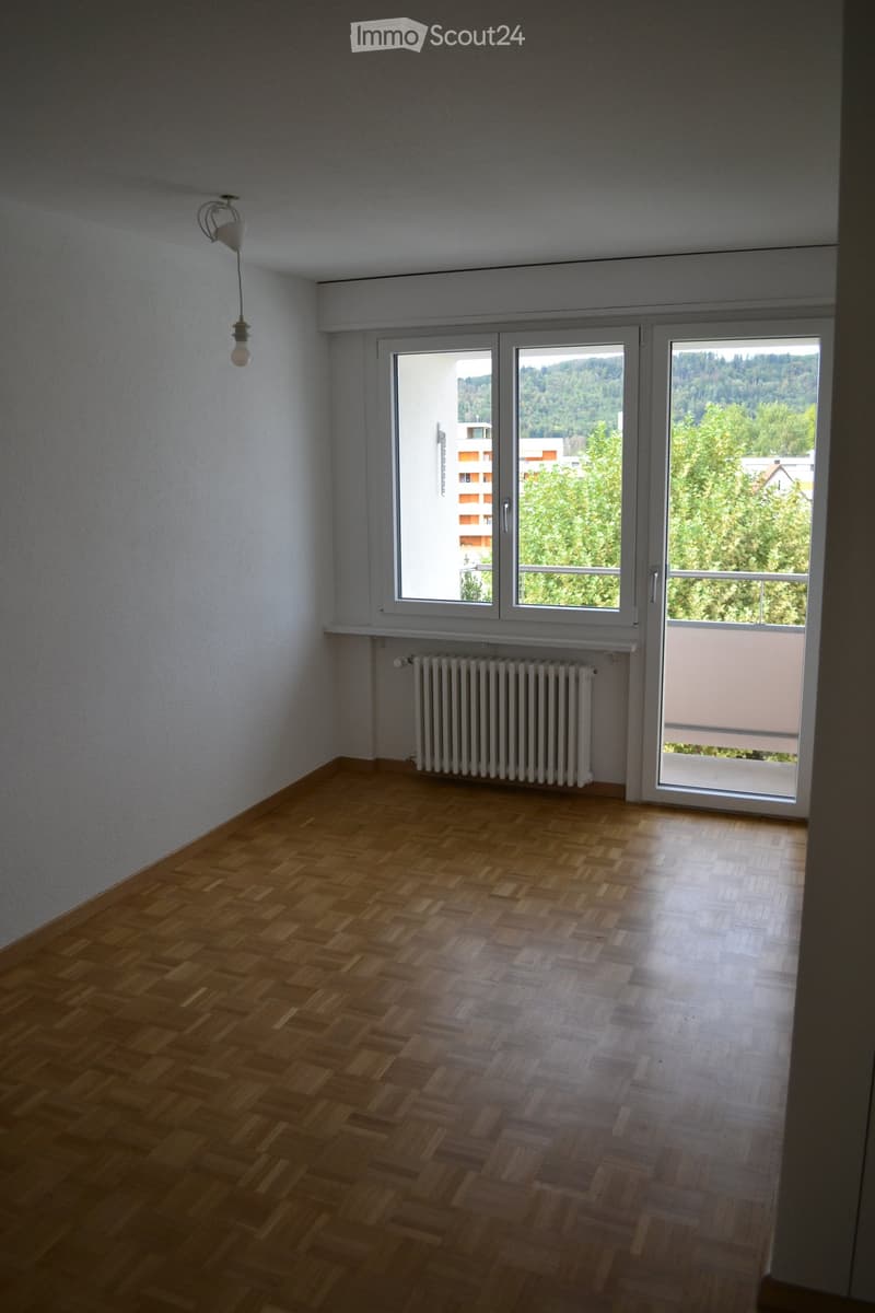 Empty room with wooden flooring, white walls, large windows, radiator, and a hanging light fixture.