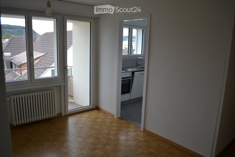 Empty living space with wooden flooring, large windows, radiator, and an adjacent kitchen area with white cabinets and appliances.