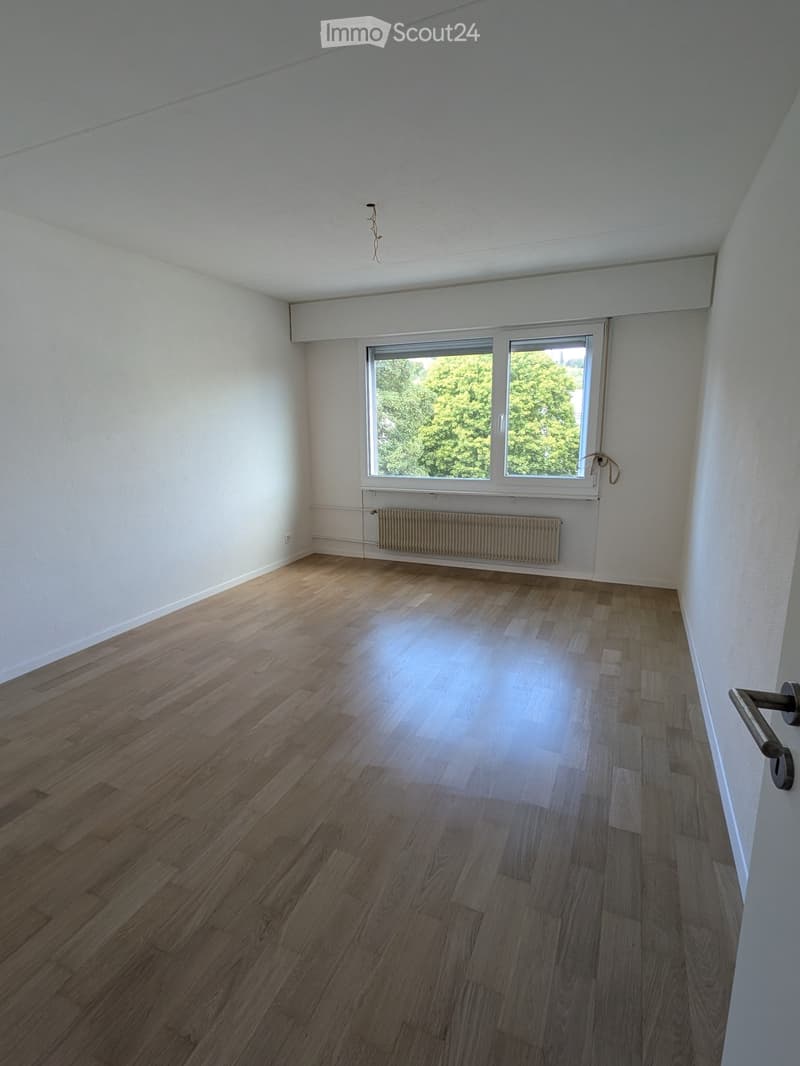 Empty room with wooden flooring, white walls, and large windows showing greenery outside. No furniture or equipment present.