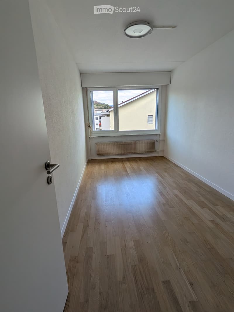 Empty room with wooden flooring, white walls, a large window with a view, a radiator, and a door with a handle.