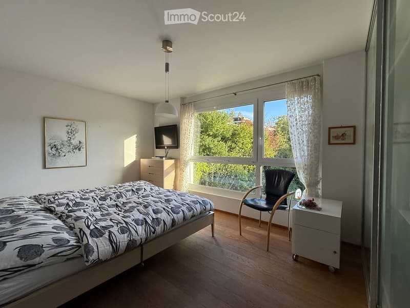 A modern bedroom with a bed, nightstand, chair, lamp, television, and picture frame. Large windows offer a view of greenery outside.