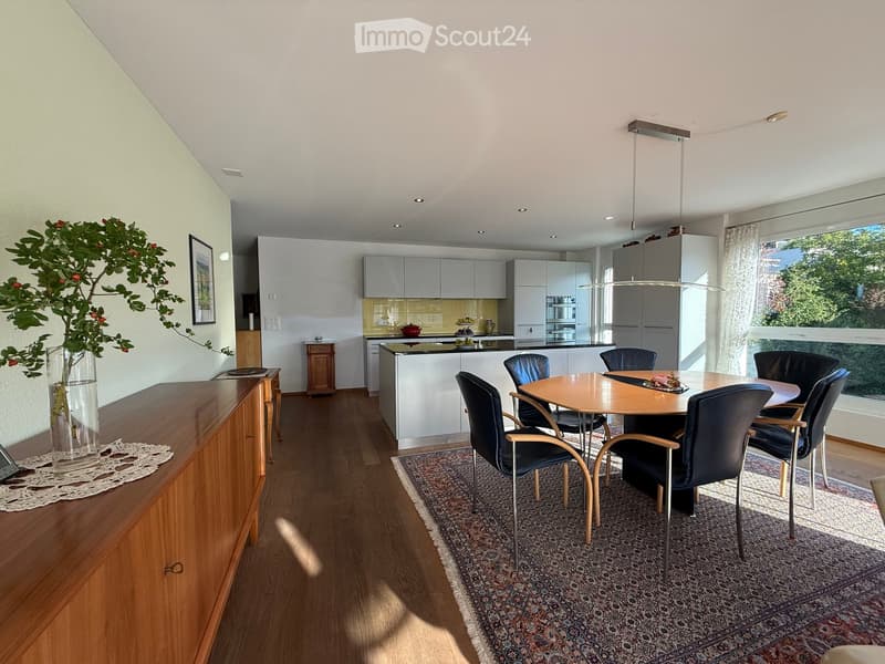 Open-plan dining and kitchen area with modern white cabinets, a central island, a round wooden dining table with six chairs, and a vase with flowers on a wooden sideboard.