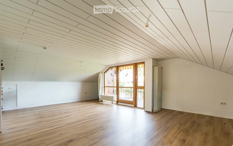 Empty attic room with wooden flooring, white walls, and a slanted ceiling. Features a large windowed door leading to a balcony or outdoor area.