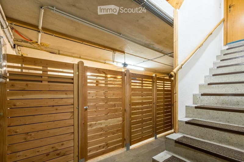 A modern hallway with a wooden gate and a staircase leading upwards, featuring exposed pipes and a skylight above.