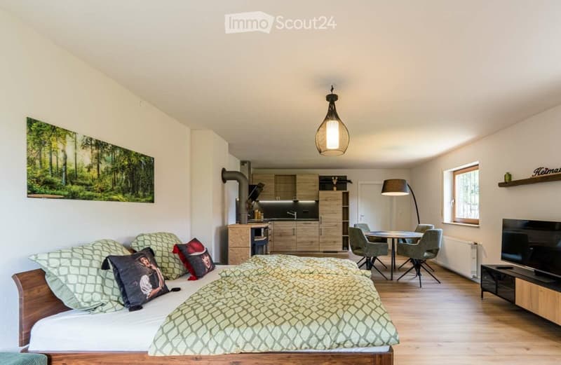 A modern, open-plan living space with a bed, kitchen area, dining table, and TV stand. The room features light wooden flooring, white walls, and contemporary furniture.