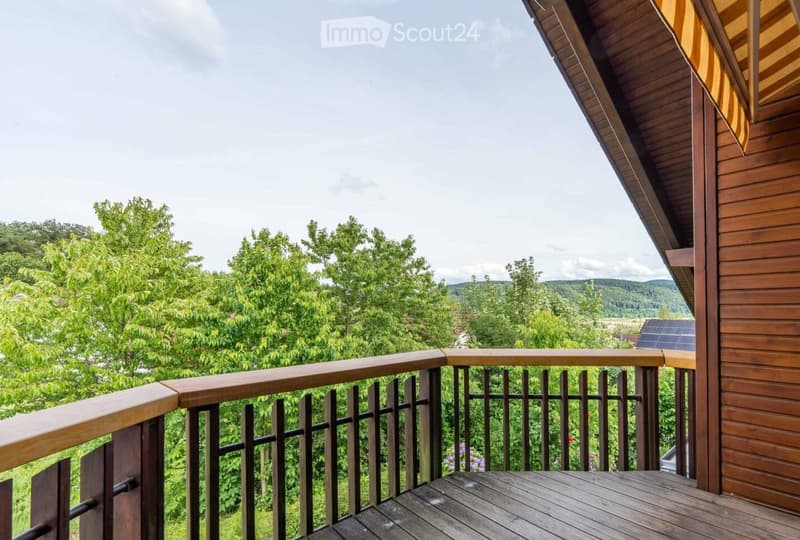 Wooden balcony with railing, overlooking a lush green landscape with trees and hills.