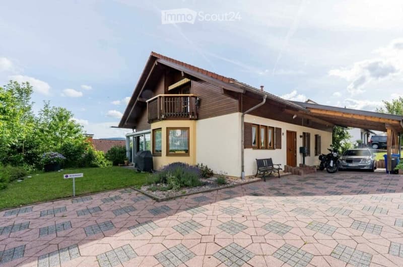Single-family house with a wooden balcony, covered entrance, and parking area for cars and motorcycles.