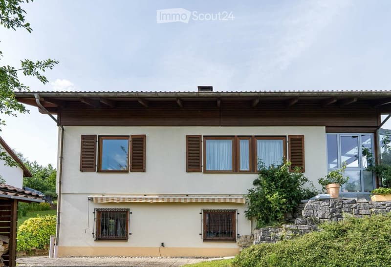 Two-story house with light beige walls, wooden shutters, and multiple windows. Features include a stone foundation, a covered entrance, and surrounding greenery.