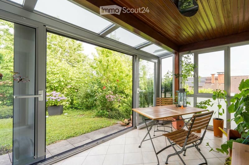 Bright sunroom with large glass walls and a wooden ceiling, featuring a wooden table, chairs, a lantern, and potted plants, overlooking a lush garden.