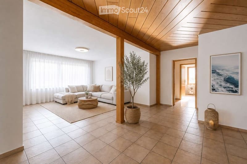 Modern living room with white walls, wooden ceiling beams, and large windows. Features a white sectional sofa, a round wicker coffee table, an ottoman, a potted plant, and decorative items.