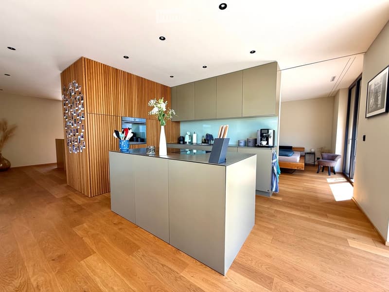 Modern kitchen with light wood flooring, grey cabinets, and a central island. Includes a coffee maker, vase with flowers, framed pictures, and a small seating area with a bed and chair.