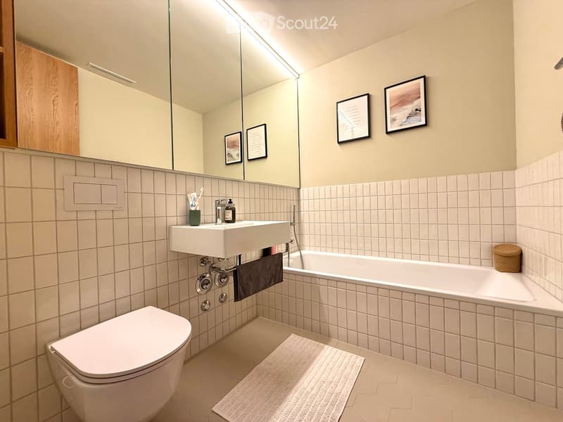 Modern bathroom with white tiles, a sink, toilet, bathtub, mirror, framed pictures, and a small woven basket on the edge of the tub.
