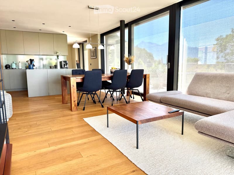 Open-plan living and dining area with wooden flooring, large windows, a sofa, coffee table, dining table with chairs, and a modern kitchen area.