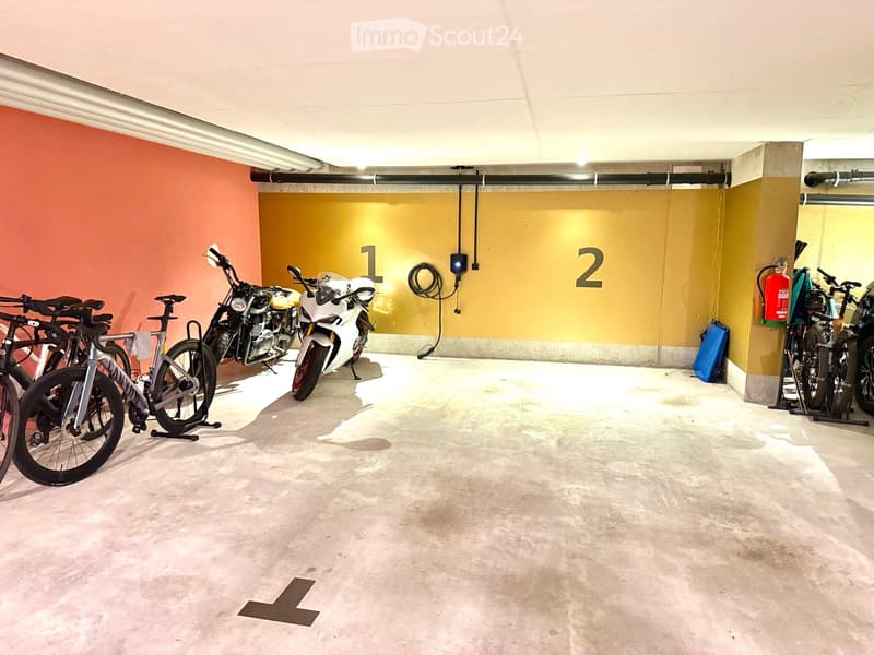 Underground parking area with several parked bicycles and motorcycles, marked sections, and a fire extinguisher on the wall.