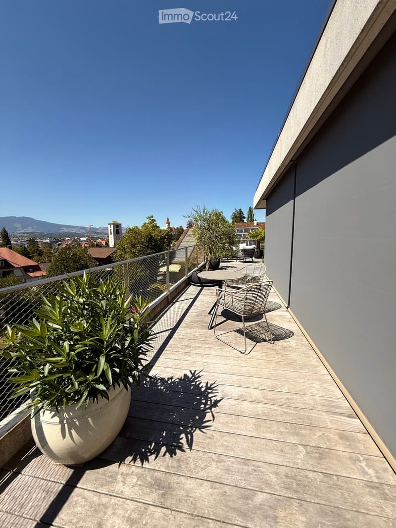 Balcony with wooden deck, metal railing, outdoor furniture, and potted plants, overlooking a cityscape with trees and buildings.