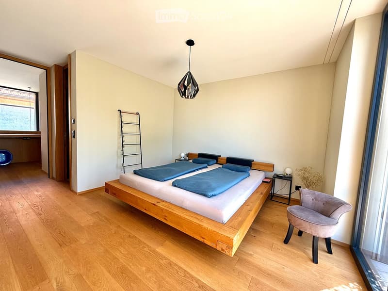 Modern bedroom with wooden flooring, a large bed with blue bedding, a grey armchair, a small table with a lamp, and a decorative plant in the corner.