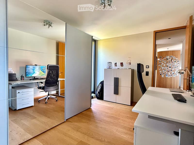 Modern office space with two desks, ergonomic chairs, computer screens, and a cabinet. The room features wooden flooring and large windows, with a glass partition separating the workspace.
