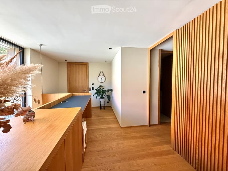 Modern hallway with light wooden flooring, white walls, and a sleek wooden bench on the left. A potted plant and a clock are visible in the background, with a doorway leading to another room on the right.