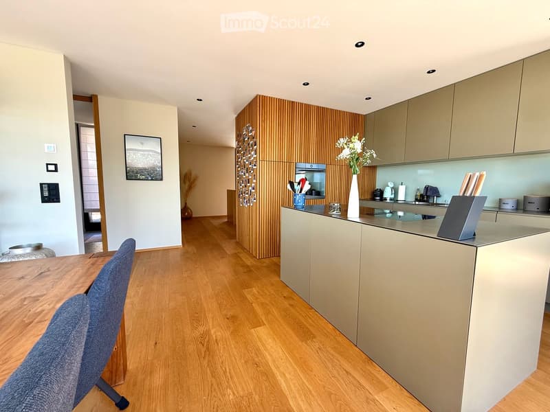 Modern kitchen with light wood flooring, light-colored cabinets, and a central island. Features include a wooden dining table with a chair, a vase with flowers, a picture frame on the wall, and various kitchen appliances.