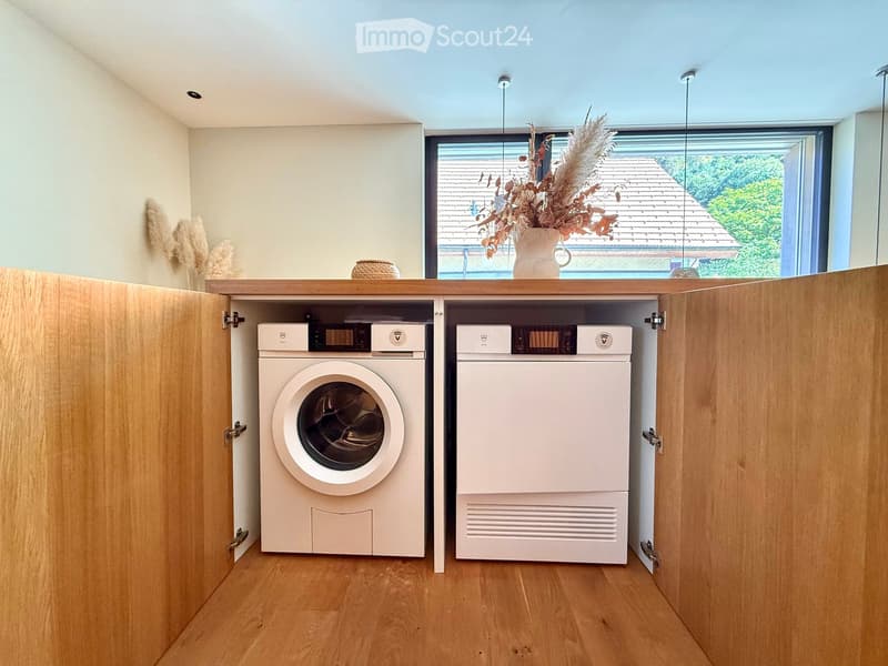 Modern bathroom with wooden cabinetry, white washing machine and dishwasher, decorative vase on windowsill, and natural light from large window.