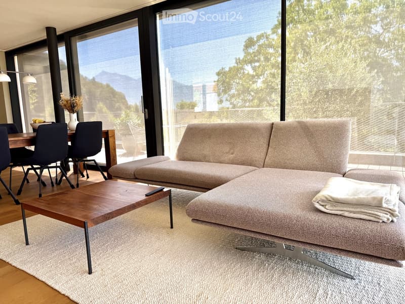 Modern living room with large windows, a sectional sofa, coffee table, dining area with chairs, and decorative vase on side table.